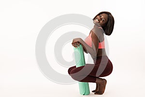 Portrait of beautiful girl in sport clothes holding a yoga mat while crouched, looking at camera and smiling, isolated
