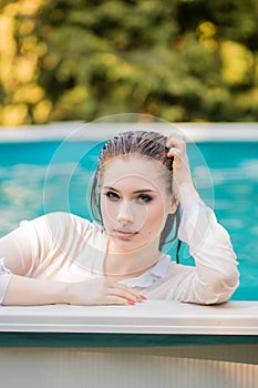 Portrait of a beautiful girl with wet hair dressed in a white shirt standing chest-deep in water near the edge of the pool