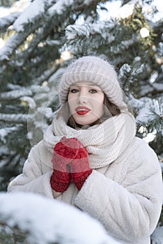 Portrait of beautiful girl in white fur coat, hat and red mittens on snow-covered fir trees background. Vertical frame