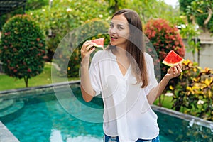 Portrait of beautiful girl in white shirt eating watermelon in courtyard of hotel near pool