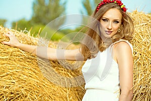 Portrait of beautiful girl in white sundress standing among haystacks at the countryside with garland made of artificial fruits on