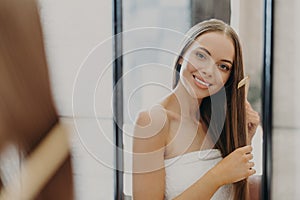 Portrait of beautiful gorgeous woman with bare shoulders combes her straight hair, smiles broadly and looks at her reflection in