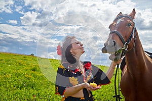 Portrait of beautiful gypsy girl with a horse on a field with green glass in summer day and blue sky and white clouds background.