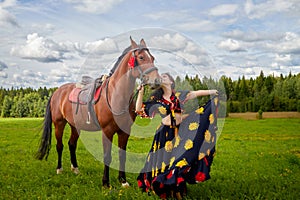 Portrait of beautiful gypsy girl with a horse on a field with green glass in summer day and blue sky and white clouds background.