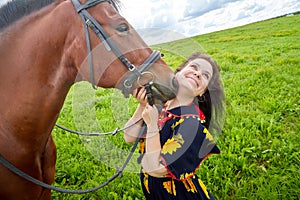 Portrait of beautiful gypsy girl with a horse on a field with green glass in summer day and blue sky and white clouds background.