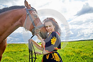 Portrait of beautiful gypsy girl with a horse on a field with green glass in summer day and blue sky and white clouds background.