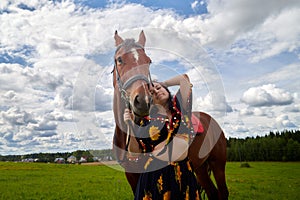 Portrait of beautiful gypsy girl with a horse on a field with green glass in summer day and blue sky and white clouds background.