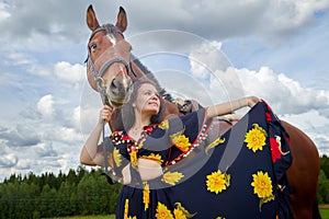 Portrait of beautiful gypsy girl with a horse on a field with green glass in summer day and blue sky and white clouds background.