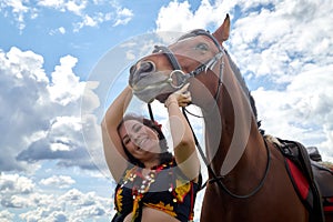 Portrait of beautiful gypsy girl with a horse on a field with green glass in summer day and blue sky and white clouds background.