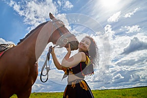 Portrait of beautiful gypsy girl with a horse on a field with green glass in summer day and blue sky and white clouds background.