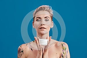 Portrait of beautiful half naked tattooed woman with short hair holding white plastic jar of cream or body lotion