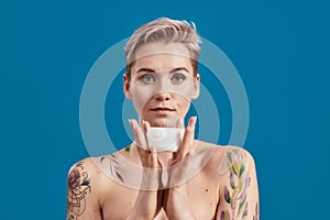 Portrait of beautiful half naked tattooed woman with short hair holding white plastic jar of cream or body lotion