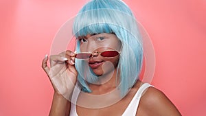 Portrait of beautiful happy Asian woman in bright blue wig and sunglasses posing over pink