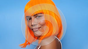 Portrait of beautiful happy Asian woman in bright orange wig posing over blue