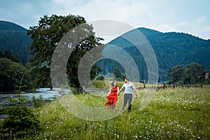 Portrait of the beautiful happy couple holding hands and walking along daisy meadow near the river in the mountains