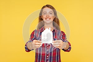 Portrait of beautiful happy ginger girl in checkered shirt showing paper house to camera and smiling, dreaming of own