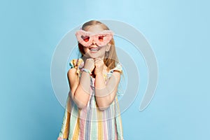 Portrait of beautiful, happy, little girl, child posing in pink furry glasses, smiling against blue studio background