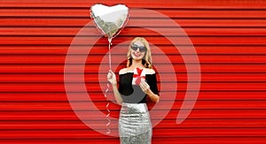 Portrait of beautiful happy smiling woman with gift box and silver heart shaped balloon on red background