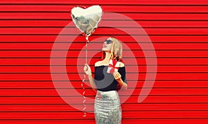 Portrait of beautiful happy smiling woman with gift box and silver heart shaped balloon on red background