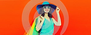 Portrait of beautiful happy smiling young woman with colorful shopping bags wearing summer straw hat on orange background, blank
