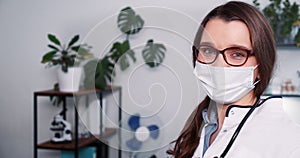 Portrait of beautiful happy young Caucasian female doctor wearing lab coat, face mask posing at clinic office workplace.