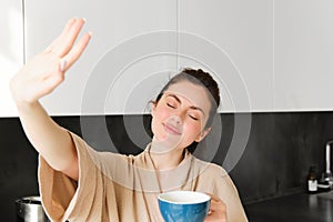 Portrait of beautiful, happy young woman covers her eyes from morning sunlight, waking up and drinking coffee, holding