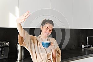 Portrait of beautiful, happy young woman covers her eyes from morning sunlight, waking up and drinking coffee, holding