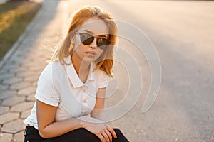 Portrait of a beautiful hipster young woman in stylish sunglasses in a white polo t-shirt in black jeans at sunset. American girl