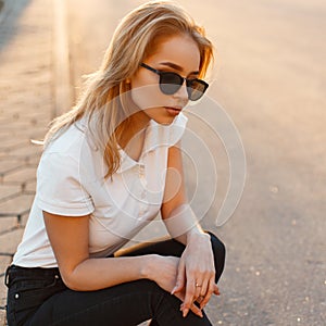 Portrait of a beautiful hipster young woman in stylish sunglasses in a white polo t-shirt in black jeans at sunset. American girl