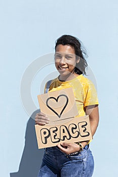 Portrait of beautiful hispanic woman with tender gesture holding a banner with text Peace. She is isolated on blue background