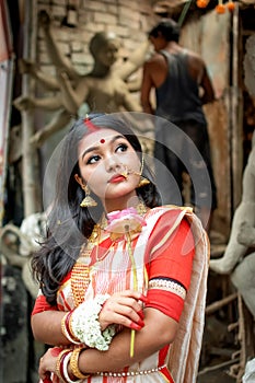 Portrait of beautiful Indian girl standing in front of Durga Idol wearing traditional Indian saree, gold jewellery, and bangles.