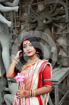 Portrait of beautiful Indian girl standing in front of Durga Idol wearing traditional Indian saree, gold jewellery, and bangles.