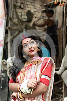 Portrait of beautiful Indian girl standing in front of Durga Idol wearing traditional Indian saree, gold jewellery, and bangles.