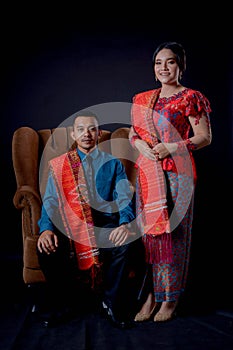 Portrait of beautiful indonesian couple wearing traditional batak costume sit on brown couch isolated on black background