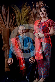 Portrait of beautiful indonesian couple wearing traditional batak costume sit on brown couch isolated on black background