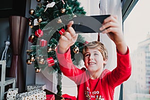portrait of beautiful kid boy at home by the christmas tree and using mobile phone to take a selfie