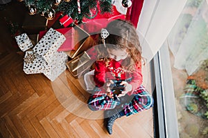 portrait of beautiful kid girl at home by the christmas tree and using mobile phone