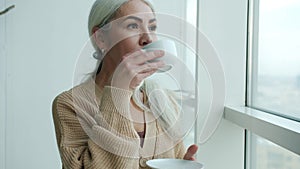 Portrait of beautiful lady wearing elegant clothing drinking tea and watching city from big window