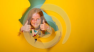 Portrait of beautiful little ginger kid girl giving you thumbs up over yellow background. breaking through yellow paper