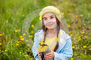 Portrait of a beautiful little girl with two pigtails holds a bouquet of yellow dandelions in a metal mug, looks up
