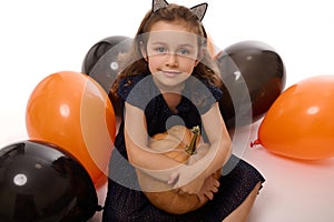 Portrait of a beautiful little girl wearing a hoop with cat ears, dressed in dark witch carnival costume, hugs a pumpkin in her