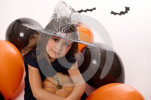 Portrait of a beautiful little girl wearing a wizard hat, dressed in dark witch carnival costume, hugs a pumpkin in her hand, sit