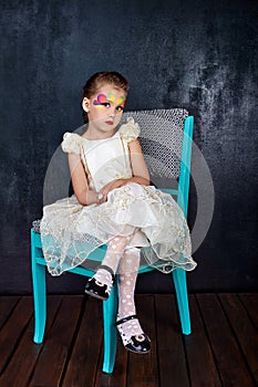 Portrait of Beautiful little girl in white dress red lips with painted face sitting on a chair at dark background