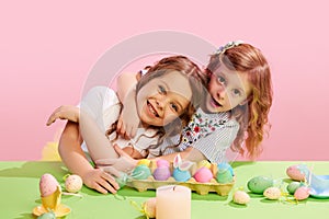 Portrait of beautiful little girls sitting at table with decorated, painted eggs, hugging, smiling against pink