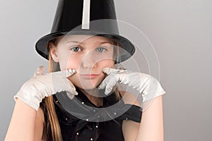 Portrait of beautiful little sad blonde girl in hat and gloves depicting smile sitting at table on gray background