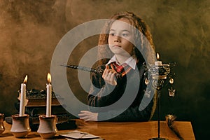 Little witch dressed in dark clothes sitting at the table against a black smoky background. There are magic wand, books