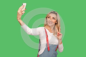 Portrait of beautiful lovely adult woman with friendly smile taking selfie on smartphone and showing victory gesture