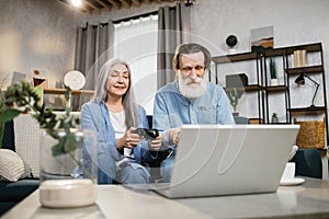 Portrait of beautiful mature woman and her bearded husband sitting on sofa and playing video games