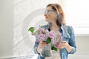 Portrait of beautiful mature woman at home with bouquet of lilac flowers. Background of home interior dining near the window