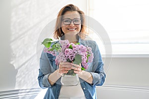 Portrait of beautiful mature woman at home with bouquet of lilac flowers. Background of home interior dining near the window